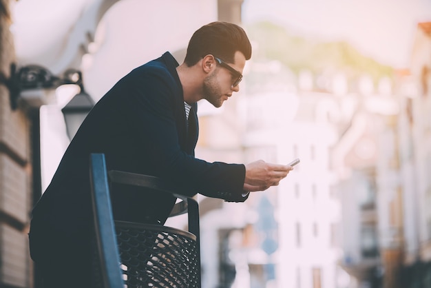 Typing message to friend. Handsome young man in smart casual wear holding smart phone and looking at it while standing outdoors