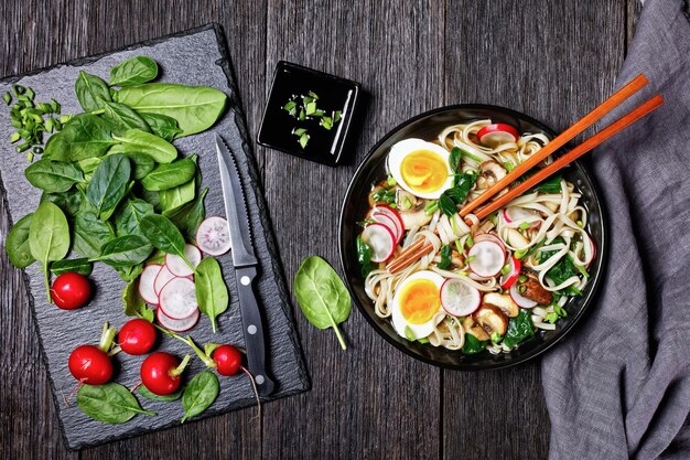 Zuppa di noodle udon - zuppa calda giapponese in una ciotola nera con bacchette in un brodo marrone con spinaci appassiti, ravanelli, uova sode, funghi affettati su uno sfondo di legno scuro, vista dall'alto, primo piano