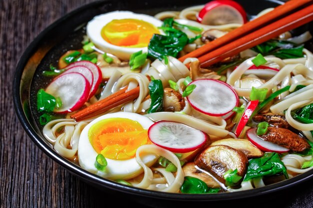 Zuppa di noodle udon - zuppa calda giapponese in una ciotola nera con bacchette in un brodo marrone con spinaci appassiti, ravanelli, uova sode, funghi affettati su uno sfondo di legno scuro, vista dall'alto, primo piano