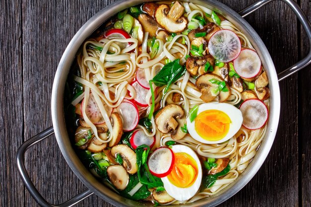 Udon noodle soup - on a metal pot of udon noodles in a broth with spinach, radish, soft boiled eggs, sliced mushrooms served on a pot on a dark wooden background, top view, close-up