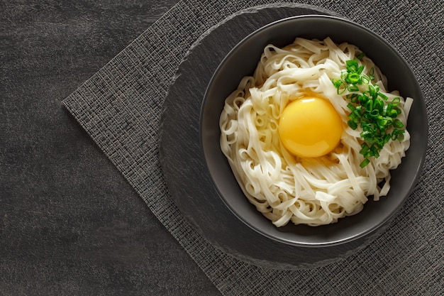 Udon noodles cooked with raw egg and green onion in a gray bowl on slate stone plate round dark background top view