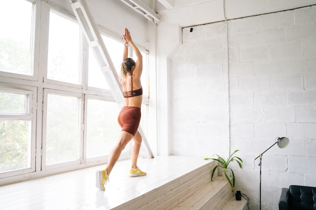 UFA, RUSSIA - 15 MAY 2020. Flexible young woman with perfect athletic body wearing sportswear doing front forward one leg step lunge exercises standing near window. Concept of healthy lifestyle
