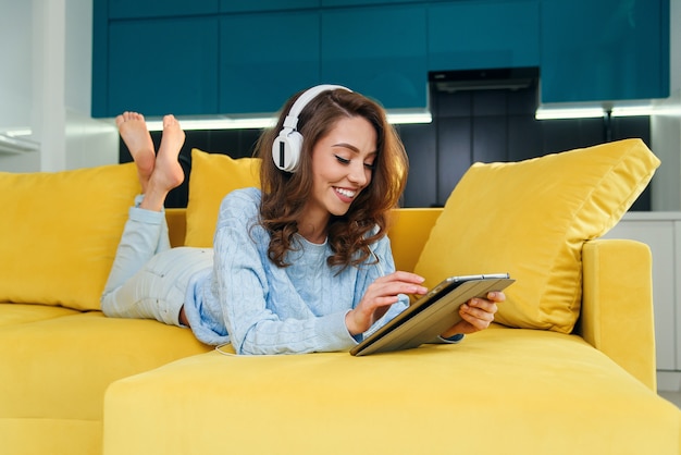 Uitbundig meisje gebruikt tablet-pc terwijl ze op de comfortabele gele bank ligt en geniet van mooie melodieën met oortelefoons.