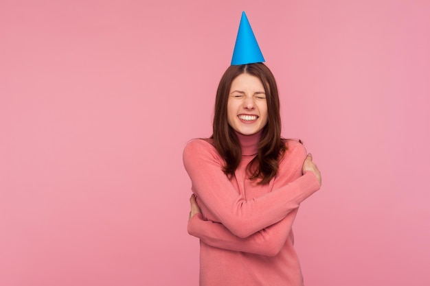 Uiterst gelukkige tevreden vrouw in roze trui met feestmuts op het hoofd die zichzelf omhelst en feliciteert met verjaardag, opgewonden met feest. Indoor studio-opname geïsoleerd op roze background