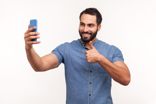 Uiterst tevreden gelukkig bebaarde man duimen opdagen en glimlachend kijken naar de camera van zijn smartphone bloggen Indoor studio shot geïsoleerd op witte achtergrond