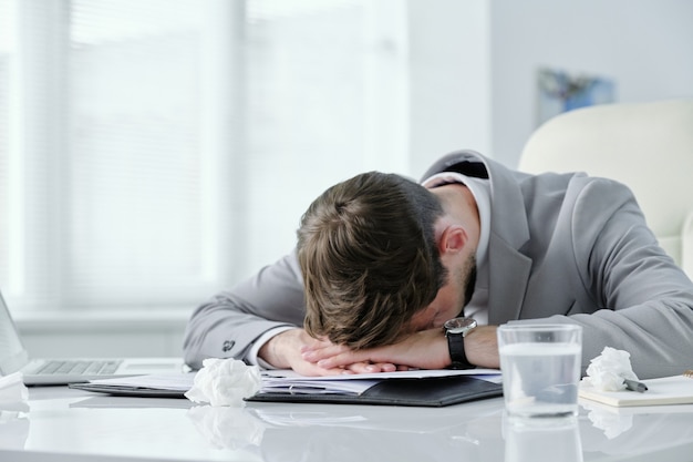 Uitgeput jonge zakenman liggend op tafel met papieren en verfrommelde servetten terwijl hij lijdt aan hoofdpijn