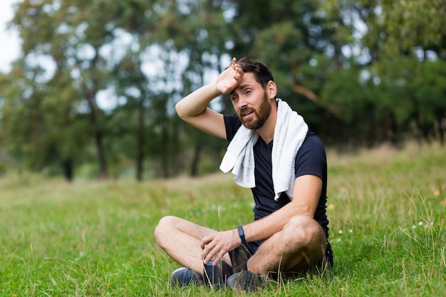 Uitgeput na training of joggen atleet man buiten zitten met een handdoek. Portret zweterige fitness man in stadspark achtergrond bos en bomen. Kaukasische sportman rust na het hardlopen