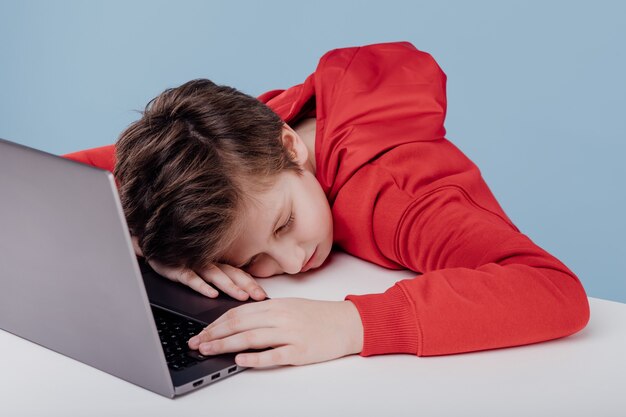 Uitgeput schooljongen dutten op tafel in de buurt van laptop in studio op blauwe achtergrond