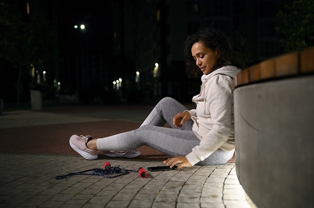 Uitgeputte atleet zit op de grond, leunend op een stenen bank, vegen op een mobiele applicatie op een smartphone tegen de achtergrond van verlicht stadsgebouw