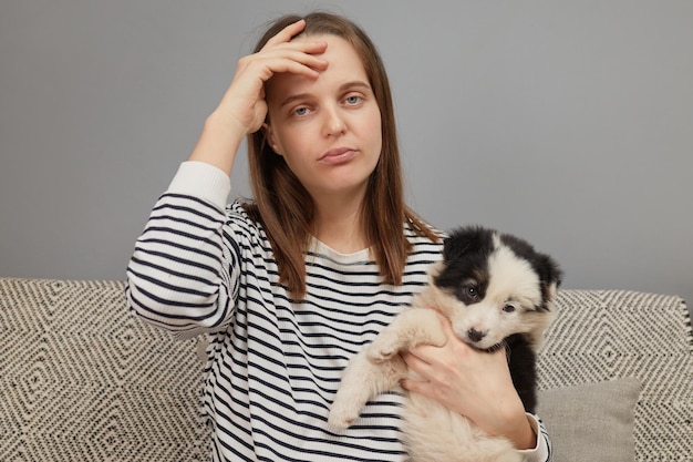 Uitgeputte blanke vrouw met een gestreepte hemd zit op de bank met een kleine hond die met banden speelt.