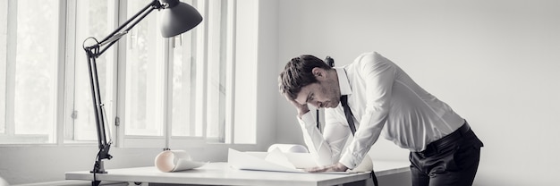 Uitgeputte en gefrustreerde zakenman die plannen of blauwdrukken analyseert aan het bureau in een modern kantoor.