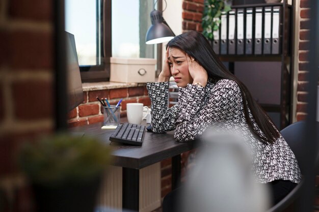 Uitgeputte kantoormedewerker met migrainehoofdpijn die aan een bureau in de werkruimte zit terwijl hij lijdt aan overwerk. Vermoeide zakenvrouw met pijnlijke hoofdpijn die zich zorgen maakt over de presentatiedeadline.