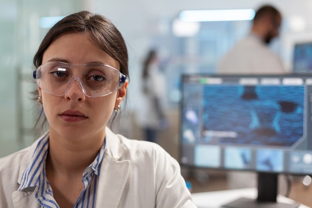 Uitgeputte microbioloog met beschermingsglas zittend in laboratorium kijkend naar camera