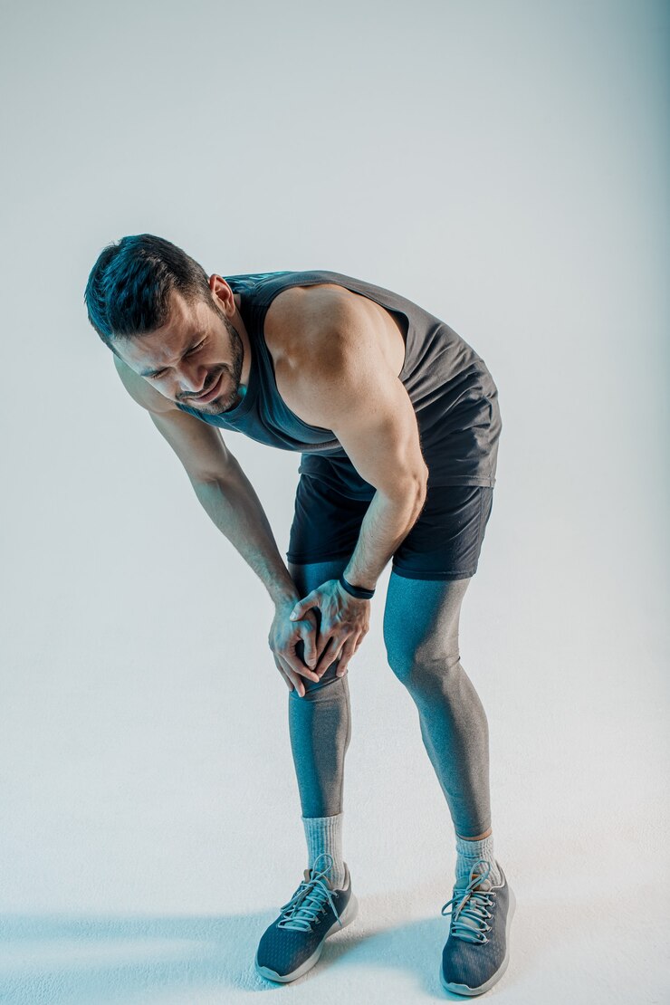 Foto uitgeputte sportman die pijn in de knie voelt. jonge, bebaarde europese man draagt sportuniform. concept van sportblessure. geïsoleerd op turkooizen achtergrond. studio shoot