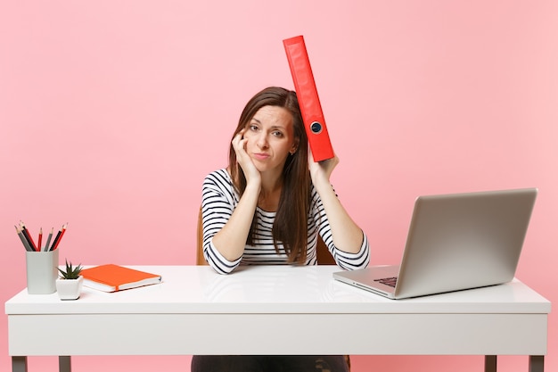 Uitgeputte vrouw met rode map met papieren document in de buurt van hoofd die aan een project werkt terwijl ze op kantoor zit met laptop