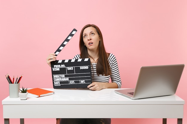 Uitgeputte vrouw met rollende ogen met klassieke zwarte film die filmklapper maakt en aan een project werkt terwijl ze op kantoor zit met een laptop