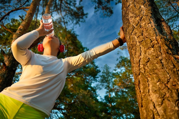 Uitgeputte vrouwelijke hardloper voelt zich niet lekker door uitdroging of overtraining Jonge dame drinkt water uit een sportfles na een zware training leunend op een boomstam