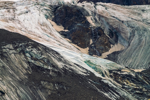 Uitlopers van gigantische gletsjer. Verbazingwekkende enorme berg rotsachtige natuurlijke muur. Rotsachtig reliëf met sneeuw en ijs in de vorm van een groot huilend oog. Diagonale besneeuwde lijn. Prachtig fantastisch kunstwerk van hooglandenaard.