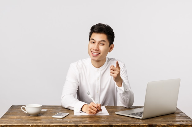 Uitstekend idee, schrijf het op. Knappe jonge Aziatische zakenman die u punt, richtend en het nemen van nota's, tevreden glimlachen, gehoord interessant idee, zittend bureau met laptop zeggen hebben