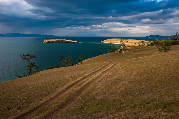 Uitzicht op het eiland aan het Baikalmeer over de steppe met een veldweg bij bewolkt weer met de zon Bomen groeien aan de rand van de kust aan de horizon er zijn bergen Zware wolken in de lucht