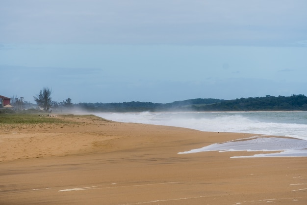 Uitzicht op het strand van Rio das Ostras in Rio de Janeiro met zonnige dag, blauwe lucht en wat wolken. Sterke zee en geelachtig zand en veel rotsen.