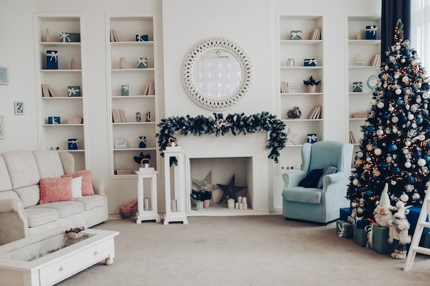 Uitzicht over witte moderne kamer ingericht voor kerstvakantie. Versierde en verlichte kerstboom, open haard met dennentak, kaarsen en handgeschept papier sterren in open haard.