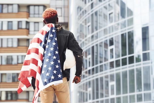 Uitzicht van achter patriot bedrijf Usa vlag conceptie van trots en vrijheid jonge afro-amerikaanse man in zwarte jas buiten in de stad staande tegen moderne zakelijke gebouw