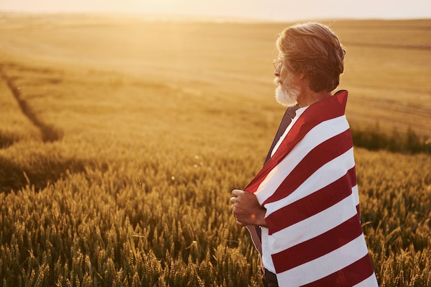 Uitzicht van achteren Holding USA vlag in handen Patriottische senior stijlvolle man met grijs haar en baard op het landbouwgebied