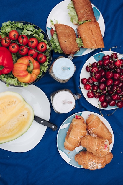 Uitzicht van bovenaf op borden met verse gezonde groenten, rode kersen, sappige meloen, heerlijke broodjes met kip, chocoladecroissants en drankjes. Concept van picknick en producten.