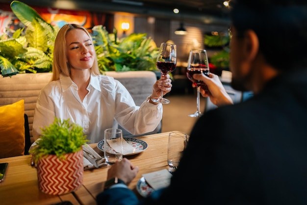 Uitzicht vanaf achterkant van onherkenbare man in pak rammelende glazen rode wijn met lachende blonde vrouw zittend aan tafel in chique restaurant 's avonds