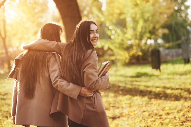 Uitzicht vanaf de achterkant van jonge lachende brunette tweeling meisjes knuffelen en plezier in casual vacht wandelen in herfst zonnig park op onscherpe achtergrond. Een van hen kijkt achteruit en houdt boeken vast.