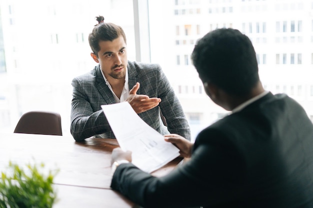 Uitzicht vanaf de achterkant van twee gerichte multi-etnische collega's die samen aan het bureau zitten in een moderne kantoorruimte en discussie over documenten, op de achtergrond van het raam. Concept van interraciale samenwerking.