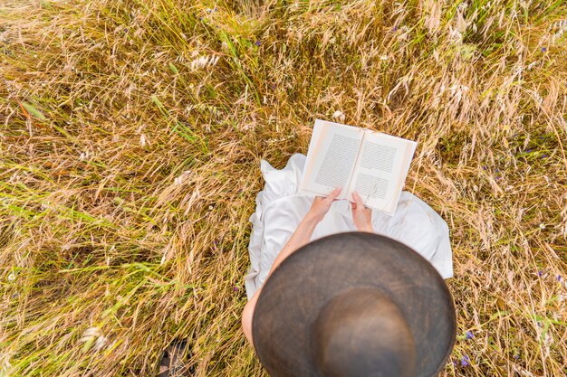 Uitzicht vanaf de top van jonge onherkenbare vrouw in witte boho-jurk zittend op een gele tarweweide die de lente viert en een boek leest. Zorgeloos meisje brengt tijd door in de natuur met haar passie voor verhalen