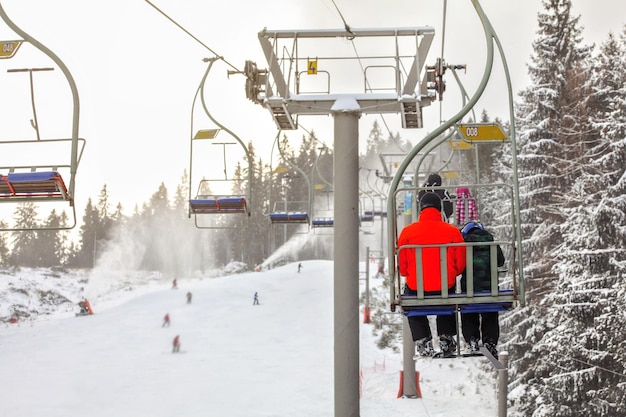 Foto uitzicht vanaf stoeltjeslift over skipiste, skiër in felrode jas vooraan, meer wazige mensen die beneden skiën, actieve sneeuwkanonnen achtergrond.