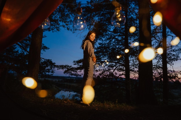 Uitzicht vanuit de tent Een jonge vrouw staat rechtop en houdt lichten in haar handen tegen de achtergrond van bomen en water Openluchtrecreatie De magie zit in de foto