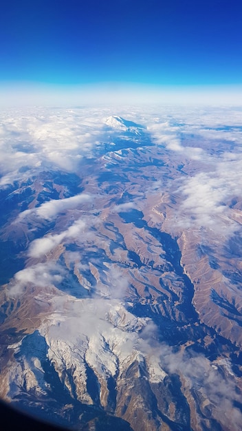 Uitzicht vanuit raam van vliegtuig naar blauwe lucht en besneeuwde bergtoppen in witte wolken