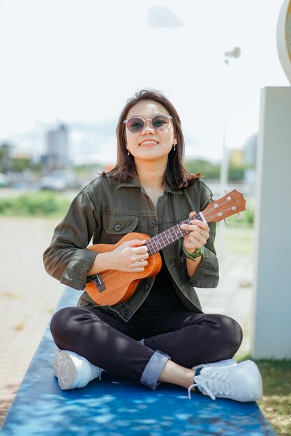 Ukelele spelen van jonge, mooie Aziatische vrouw met een jas en zwarte spijkerbroek die buitenshuis poseert