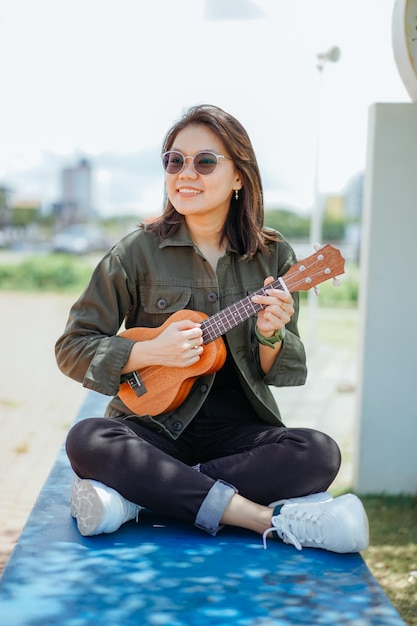 Ukelele spelen van jonge, mooie Aziatische vrouw met een jas en zwarte spijkerbroek die buitenshuis poseert