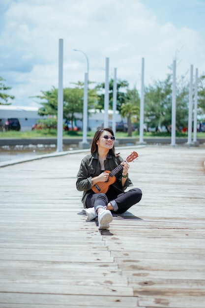 Ukelele spelen van jonge, mooie Aziatische vrouw met een jas en zwarte spijkerbroek die buitenshuis poseert