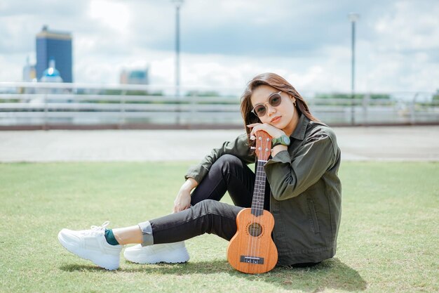 Ukelele spelen van jonge, mooie Aziatische vrouw met een jas en zwarte spijkerbroek die buitenshuis poseert