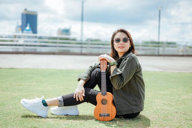 Ukelele spelen van jonge, mooie Aziatische vrouw met een jas en zwarte spijkerbroek die buitenshuis poseert