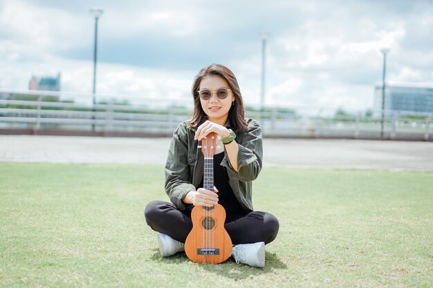 Ukelele spelen van jonge, mooie Aziatische vrouw met een jas en zwarte spijkerbroek die buitenshuis poseert