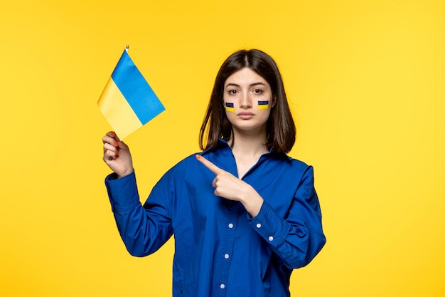 Ukraine russian conflict young pretty girl flags on cheeks yellow background pointing at flag