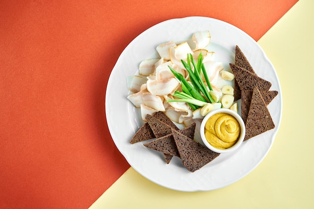 Ukrainian appetizer sliced bacon (salo) with rye bread and mustard on a white plate on bright colored surface