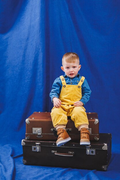 노란색 작업복을 입은 3세 우크라이나 소년은 여행가방을 가지고 노는 파란색 배경의 현대 어린이 행복한 아이