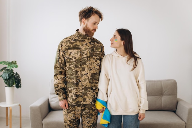 A Ukrainian couple a military man in uniform and his girlfriend are connected by a Ukrainian flag at home