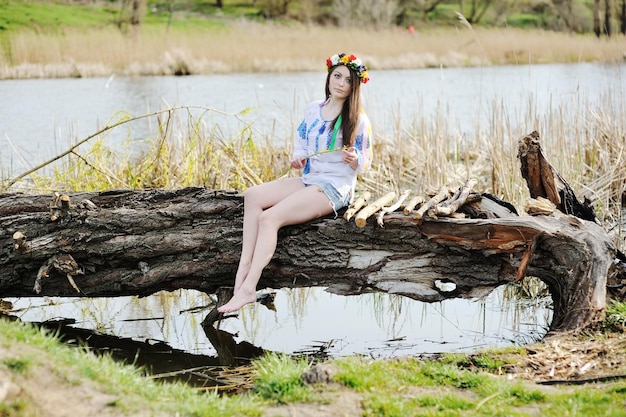 사진 드레스를 입은 우크라이나 소녀와 강가의 통나무에 앉아 머리에 꽃 화환