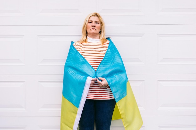 Ukrainian mature woman with blond hair serious gesture with\
blue and yellow ukrainian flag on white background in the street\
ukraine war invasion protest patriotism and no war concept