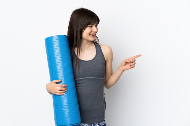 Ukrainian sport girl with mat isolated on blue wall pointing to the side to present a product