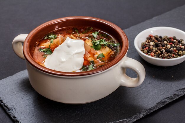 Ukrainian traditional borsch with sour cream in porcelain bowl and plate with allspice berries on stone board. Top view.